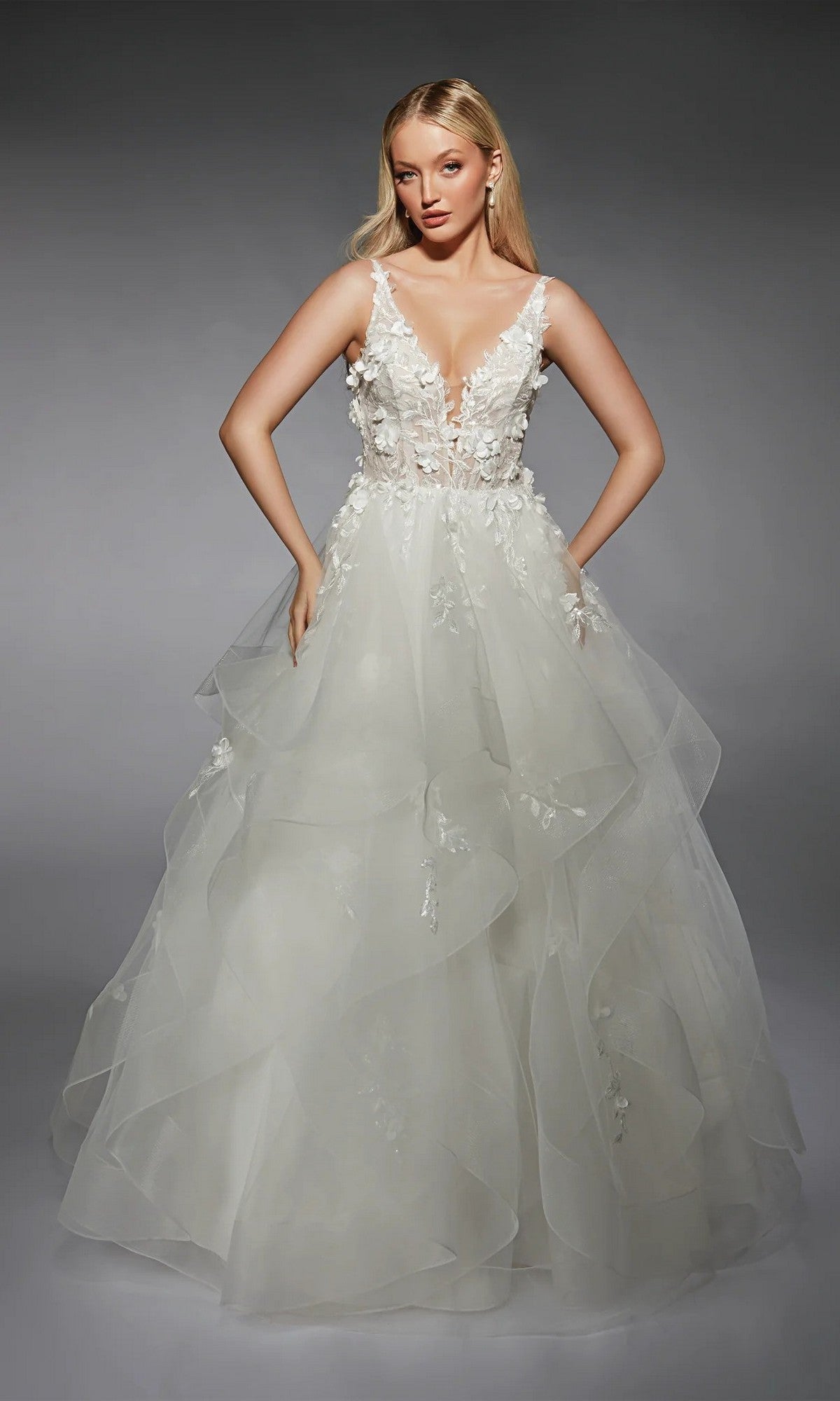 A woman wears the Alyce 7131 long white dress—sleeveless with a deep V-neck, 3D floral lace bodice, and layered ruffled tulle skirt—standing against a plain gray background.